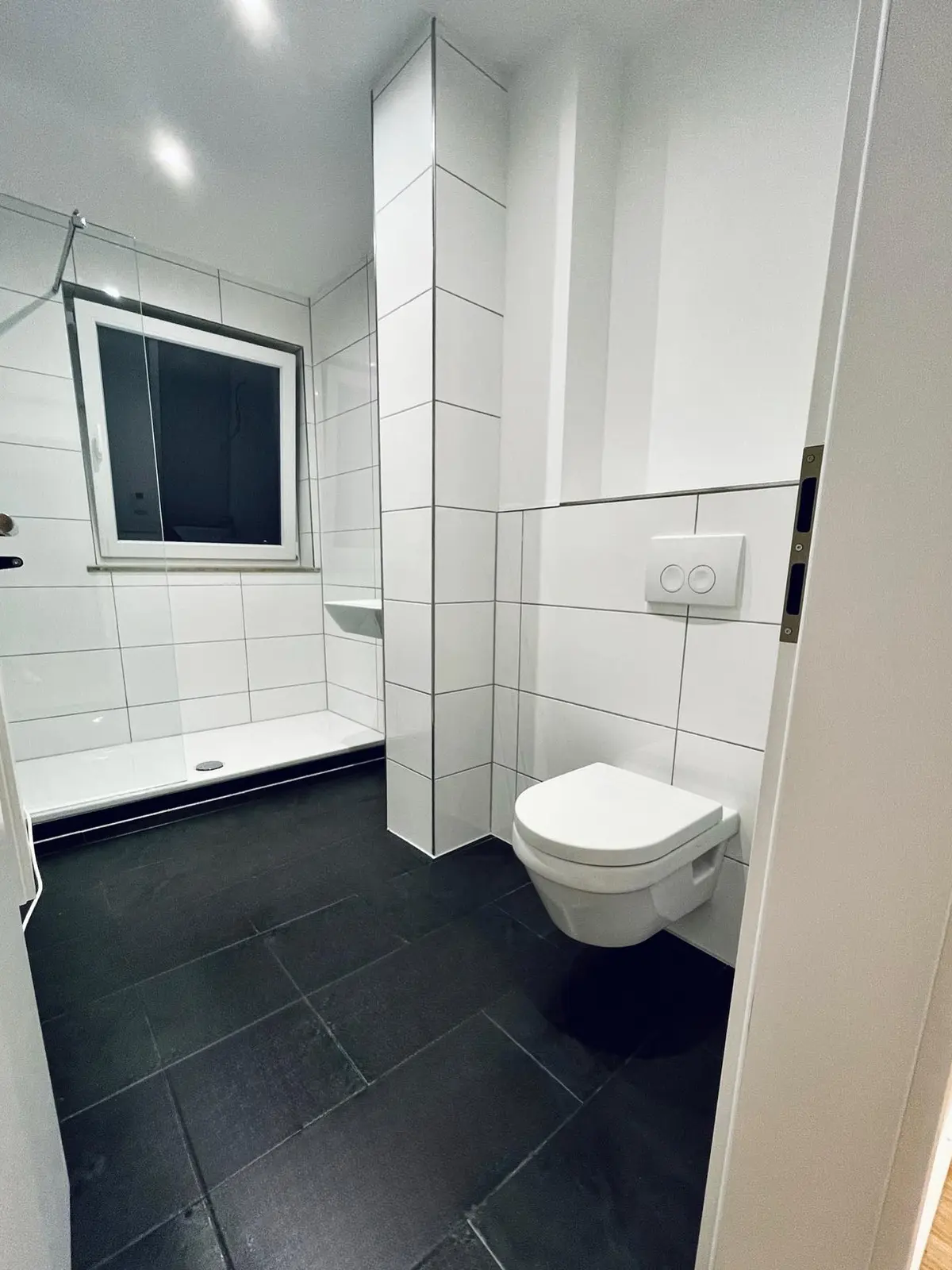 Referenz: saniertes Bad mit weißen Fliesen, schwarzem Boden und Hänge-WC nach Kernsanierung