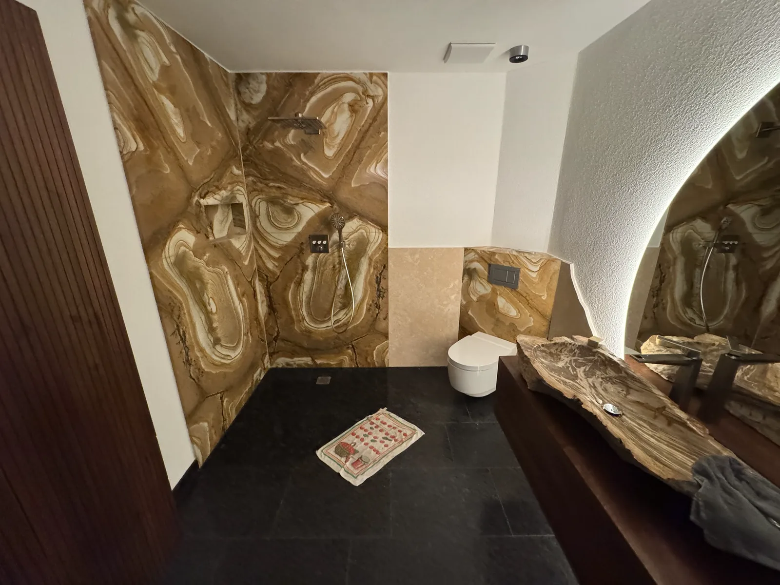 Nachher: fertiges Bad mit bodengleicher Dusche, großformatigen Wandplatten in Onyx-Optik, Wand-WC, Naturstein-Waschtisch und hinterleuchtetem Spiegel